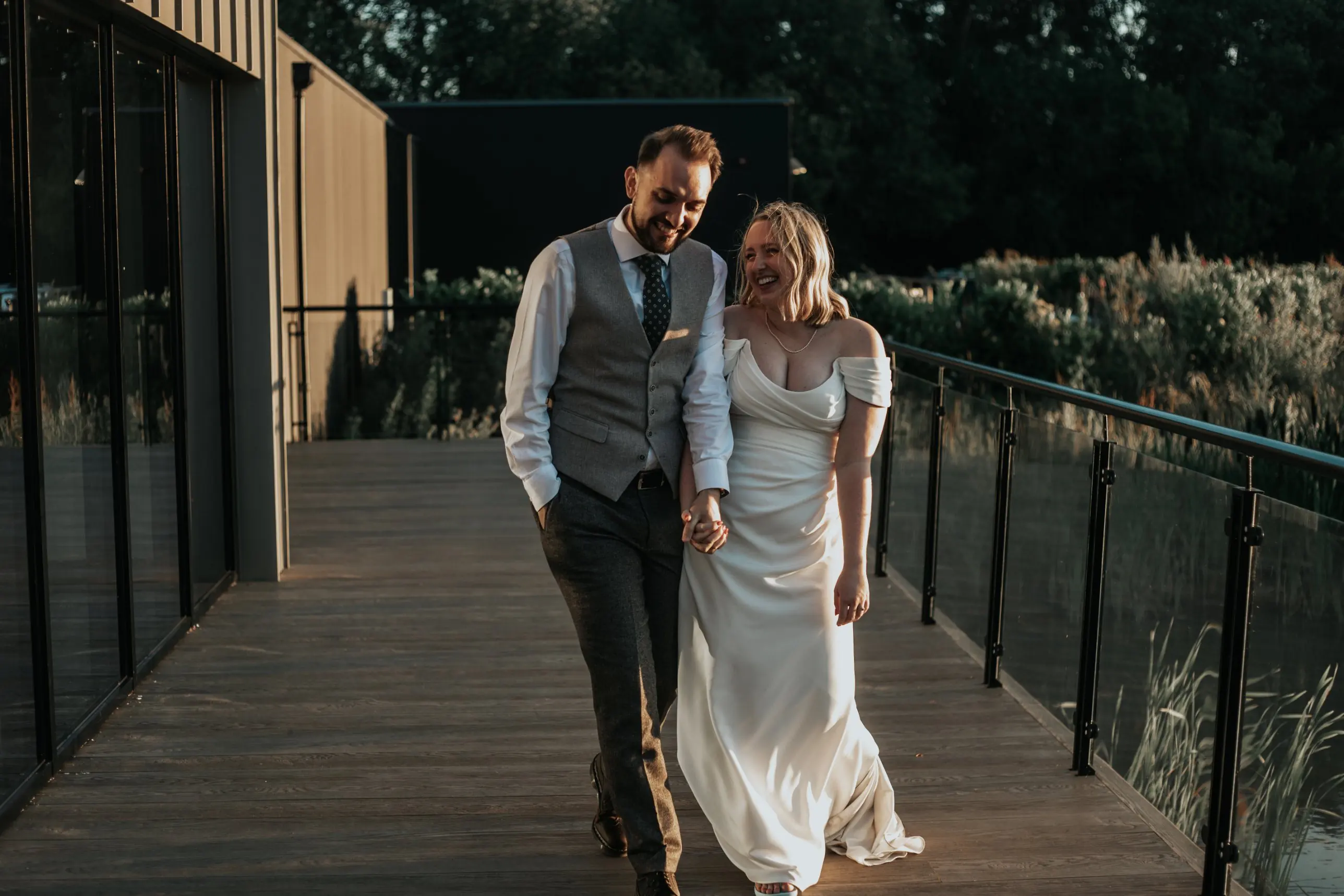 Bride and groom walking hand in hand along a lakeside deck at golden hour, laughing